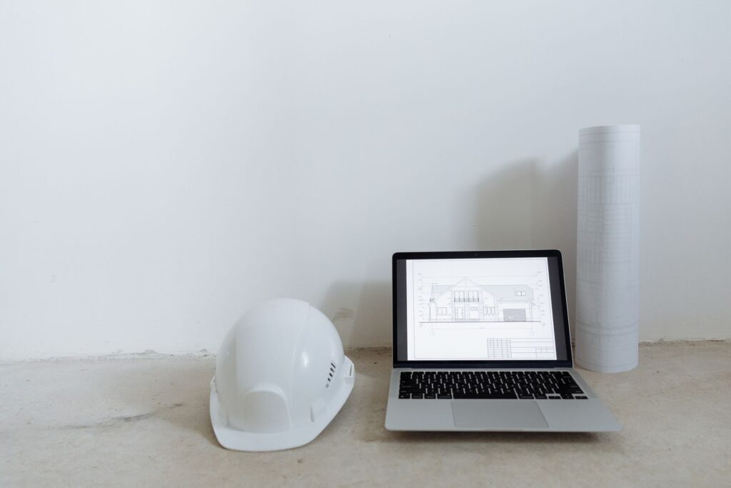 Laptop showing a house elevation drawing beside a hard hat and rolled plans, like a builder landing page